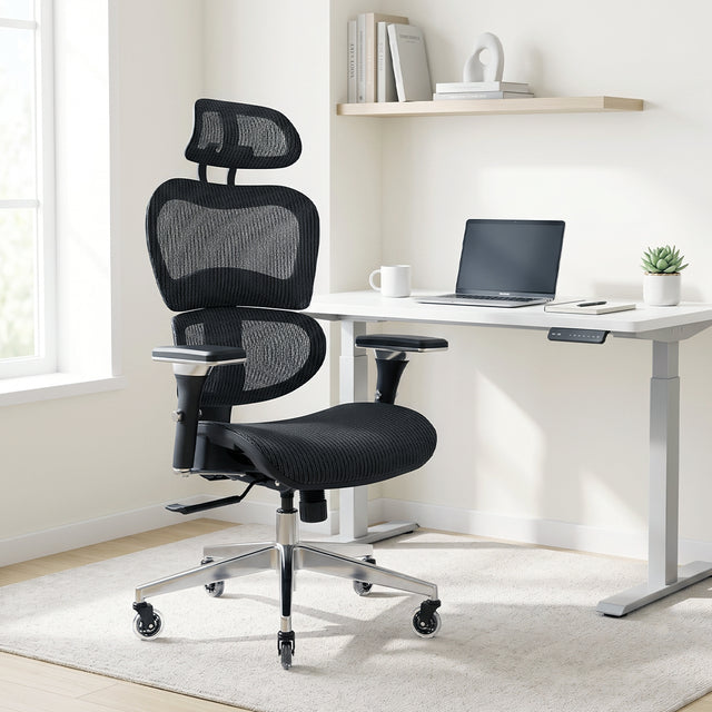 NHO-0001BL-V2, Black Coffee ergonomic mesh office chair in a bright, modern home office during a sunny afternoon. The high-back black chair features an adjustable headrest, lumbar support, and a polished silver base with casters, resting on a cream rug. In the background, natural sunlight illuminates a minimalist white standing desk with a laptop and a floating shelf with books.