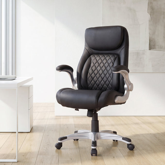 NHO-0004BL, Flat Black ergonomic executive office chair in a bright, modern home office during a sunny daylight saving time afternoon. The premium black leather chair features a diamond-quilted backrest, padded flip-up armrests, and a sleek silver base with casters, resting on light hardwood floors. In the background, warm natural sunlight illuminates a minimalist white desk setup and an abstract textured wall painting, creating a professional yet serene remote work environment.