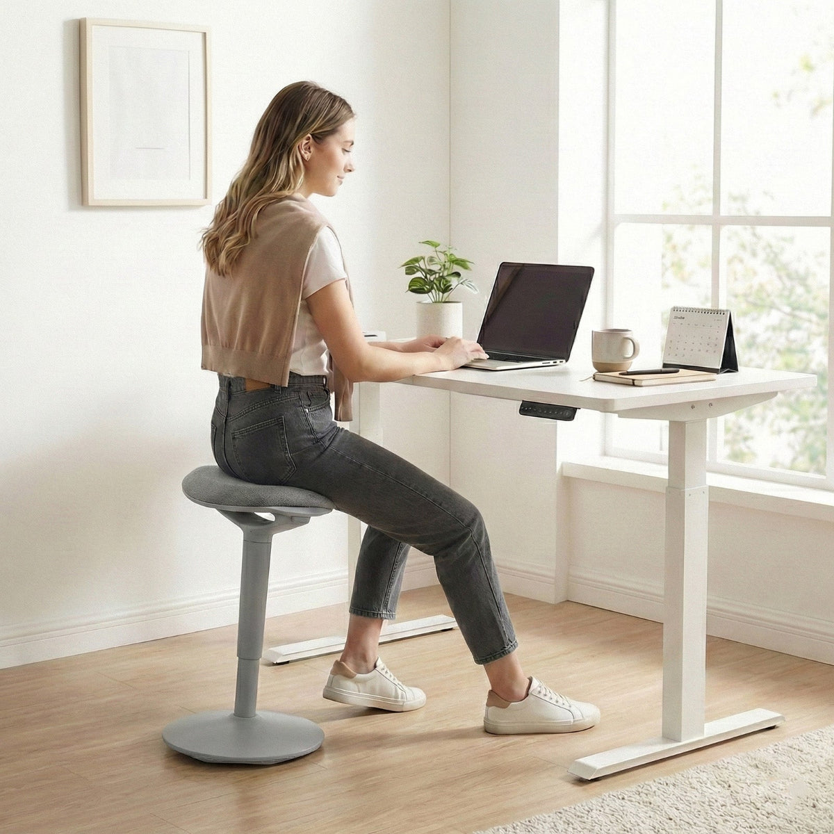 NH0-0019GR, woman using a gray active stool at a white standing desk in a bright, modern home office, demonstrating dynamic sitting posture while working on a laptop.