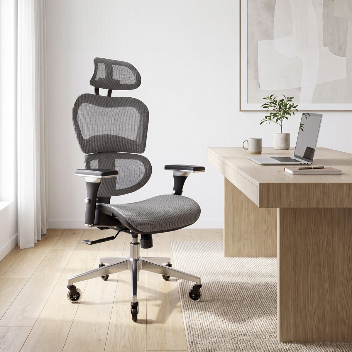 NHO-0001GR-V2, Silver Grey ergonomic mesh office chair in a bright, modern home office during a sunny afternoon. The high-back grey chair features an adjustable headrest, lumbar support, and a polished silver base with rollerblade casters, resting on a textured rug over light hardwood floors. In the background, natural sunlight illuminates a sleek wooden desk with a laptop, mug, a small potted plant, and abstract wall art.