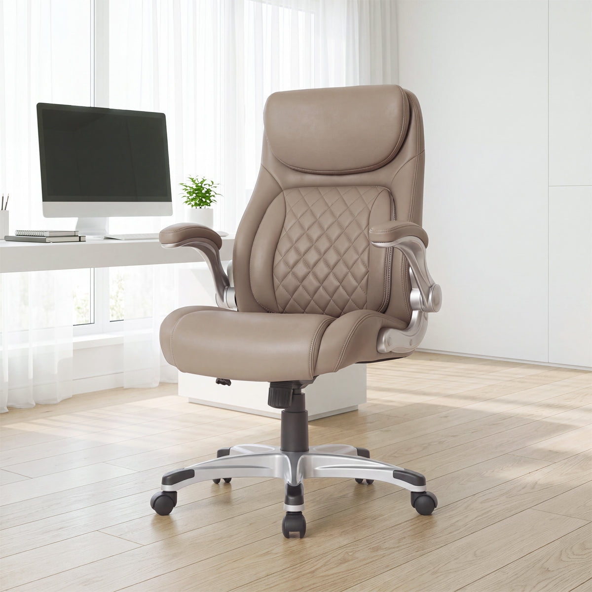 NHO-0004TP, Flat Taupe ergonomic executive office chair in a bright, modern American home office during a sunny daylight saving time afternoon. The premium taupe leather chair features a diamond-quilted backrest, padded flip-up armrests, and a sleek silver base with casters, resting on light hardwood floors. In the background, warm natural sunlight streams through sheer curtains, illuminating a minimalist white desk with a computer and a small potted plant.