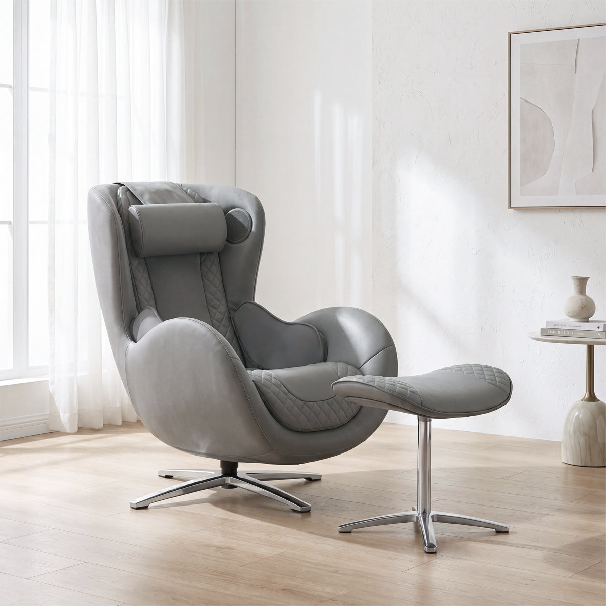 N-0003AG-V2, Ash Grey ergonomic massage armchair with matching ottoman in a bright, contemporary American home during a sunny daylight saving time afternoon. The sophisticated all-grey leather lounger features detailed diamond quilting and sits on light hardwood floors. In the background, warm natural daylight streams through a large window with sheer white curtains, casting soft shadows on the clean white walls and illuminating a sleek pedestal side table with a decorative vase and abstract wall art.