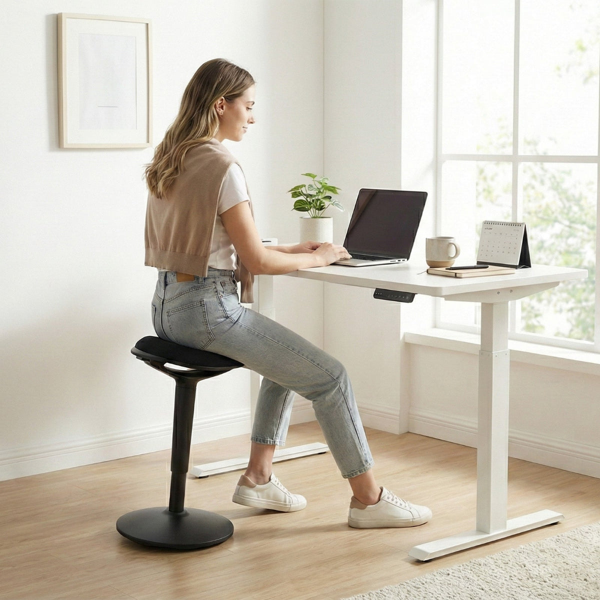 NH0-0019BL, woman using a black active stool at a white standing desk in a bright, modern home office, demonstrating dynamic sitting posture while working on a laptop.